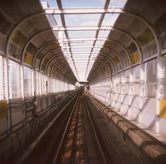 從車頭望向前方，鐵軌穿過拱形鋼架與玻璃圍成的高架軌道，頂部透出藍天。